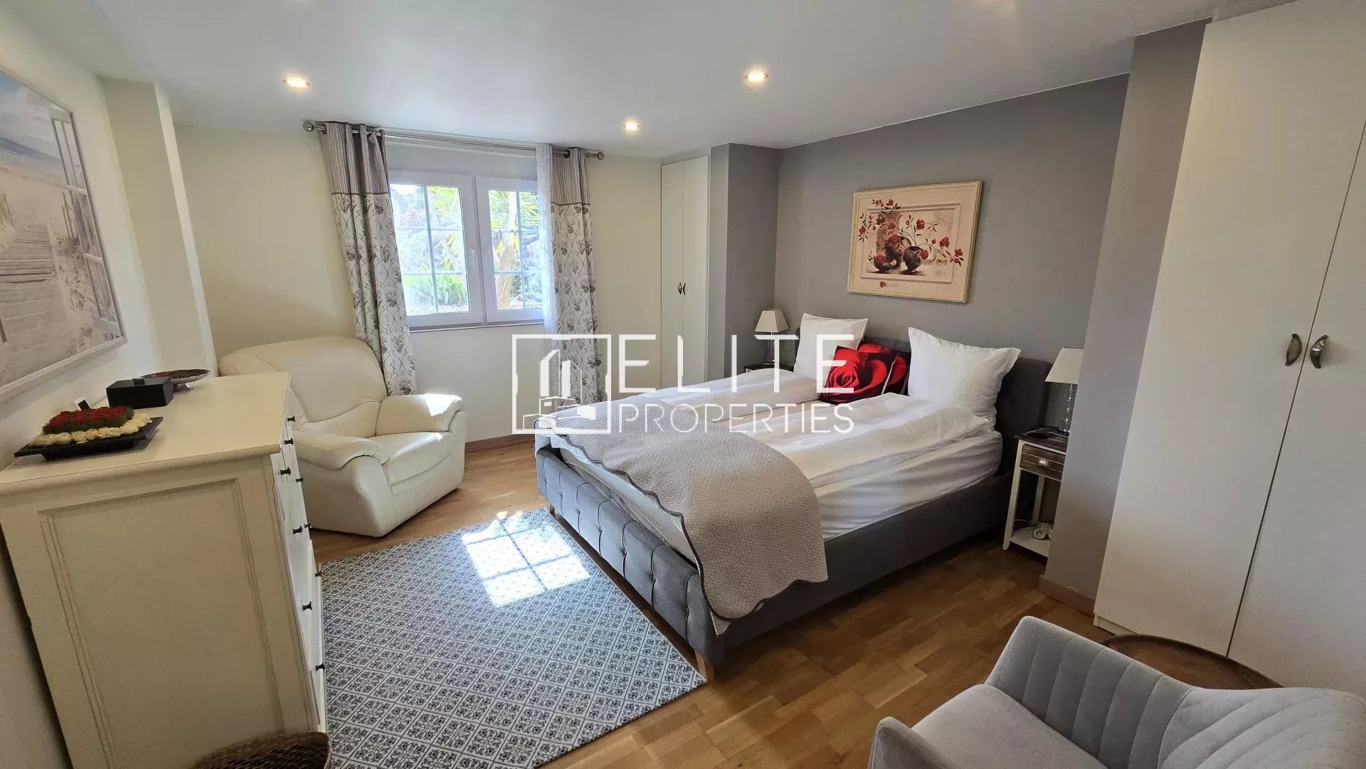Bedroom with a gray upholstered bed, white bedding, red cushions, bedside tables, and a window with patterned curtains.