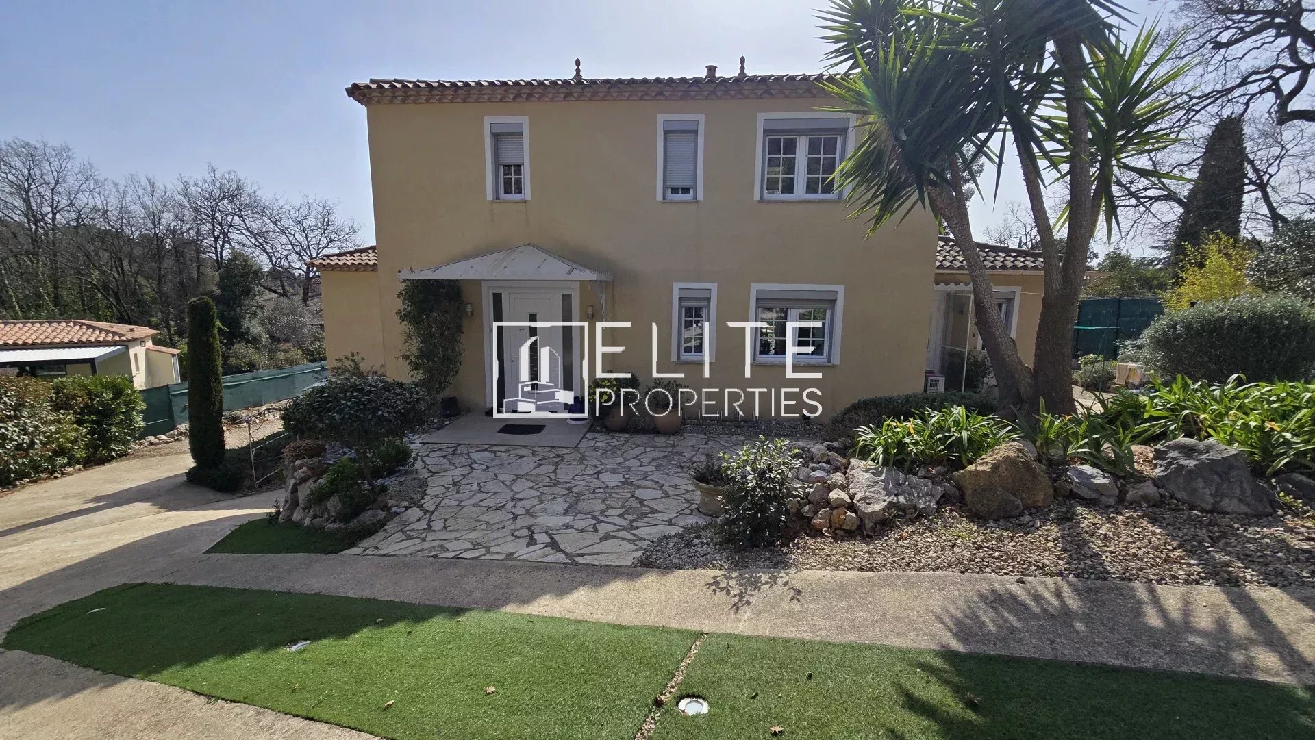 Two-story beige house with tiled roof and white-framed windows, front entrance under a small awning, stone path leading to the door, and landscaped yard.