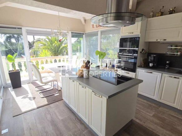Bright modern kitchen with white cabinets and a central island, opening to a sunlit patio with glass doors and lush greenery outside.