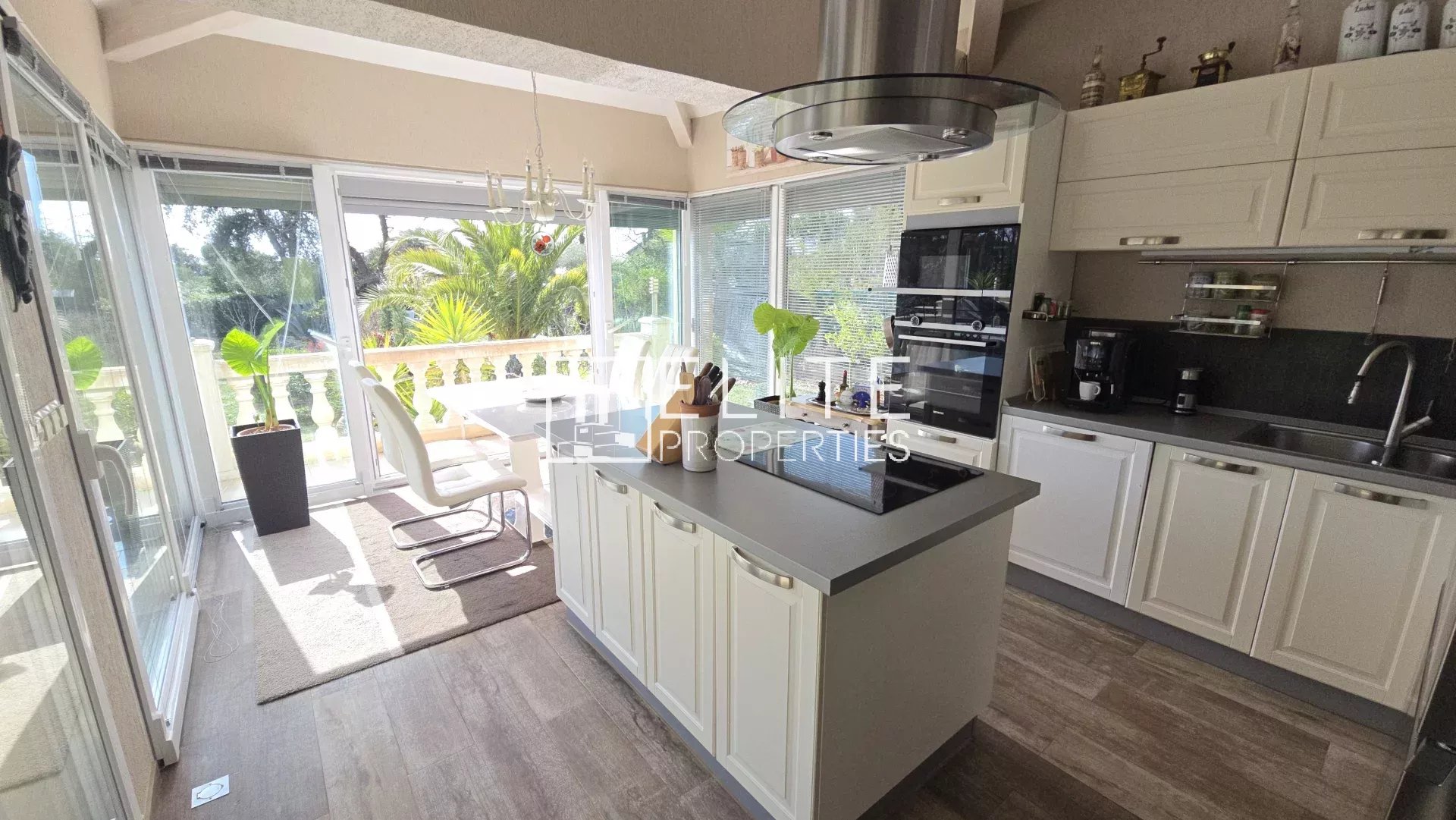 Bright modern kitchen with white cabinets and a central island, opening to a sunlit patio with glass doors and lush greenery outside.