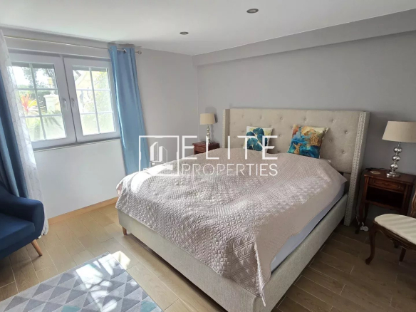 Light gray bedroom with a tufted beige bed, two wooden nightstands, matching lamps, blue curtains, and a blue armchair by the window.