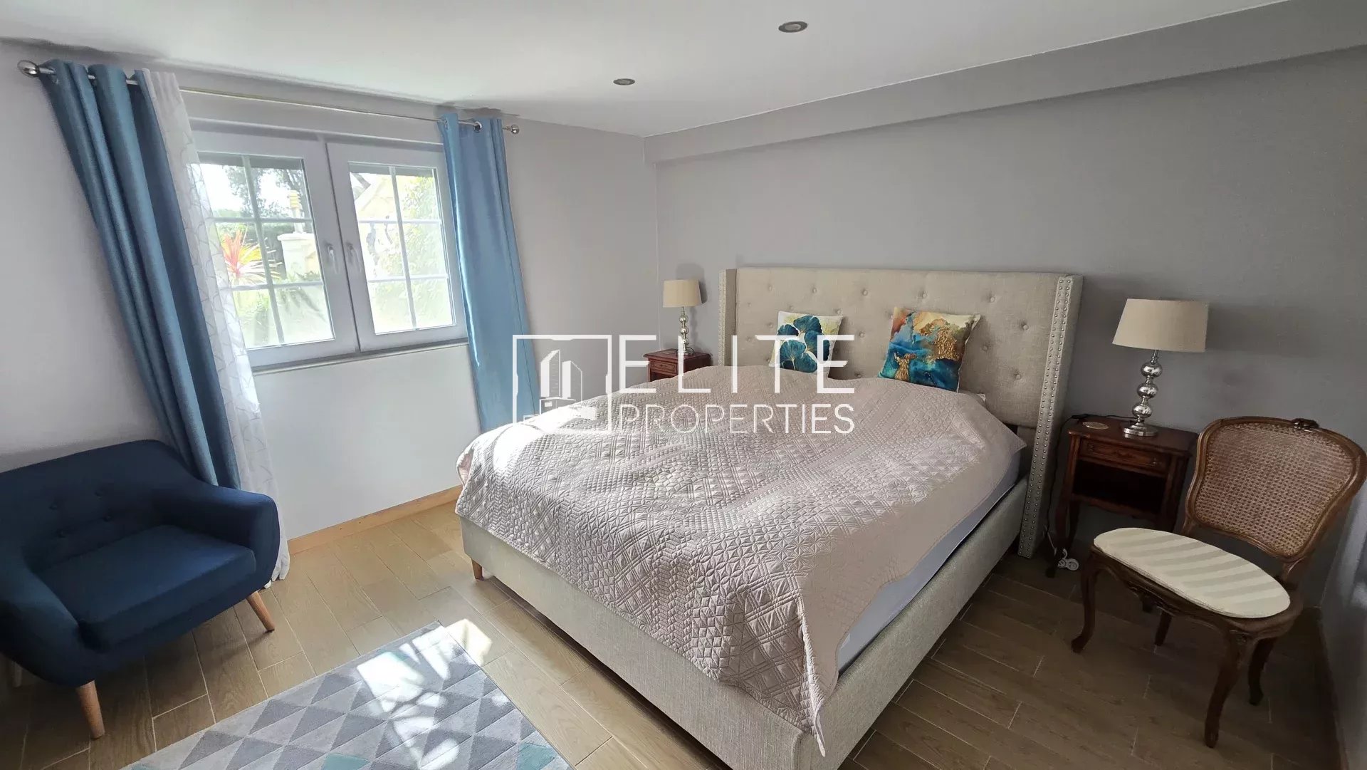 Light gray bedroom with a tufted beige bed, two wooden nightstands, matching lamps, blue curtains, and a blue armchair by the window.