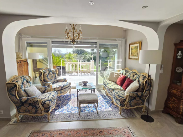 Elegant living room with blue and gold upholstered sofas facing a glass coffee table and chandelier, opening to a sunny outdoor patio through sliding doors.