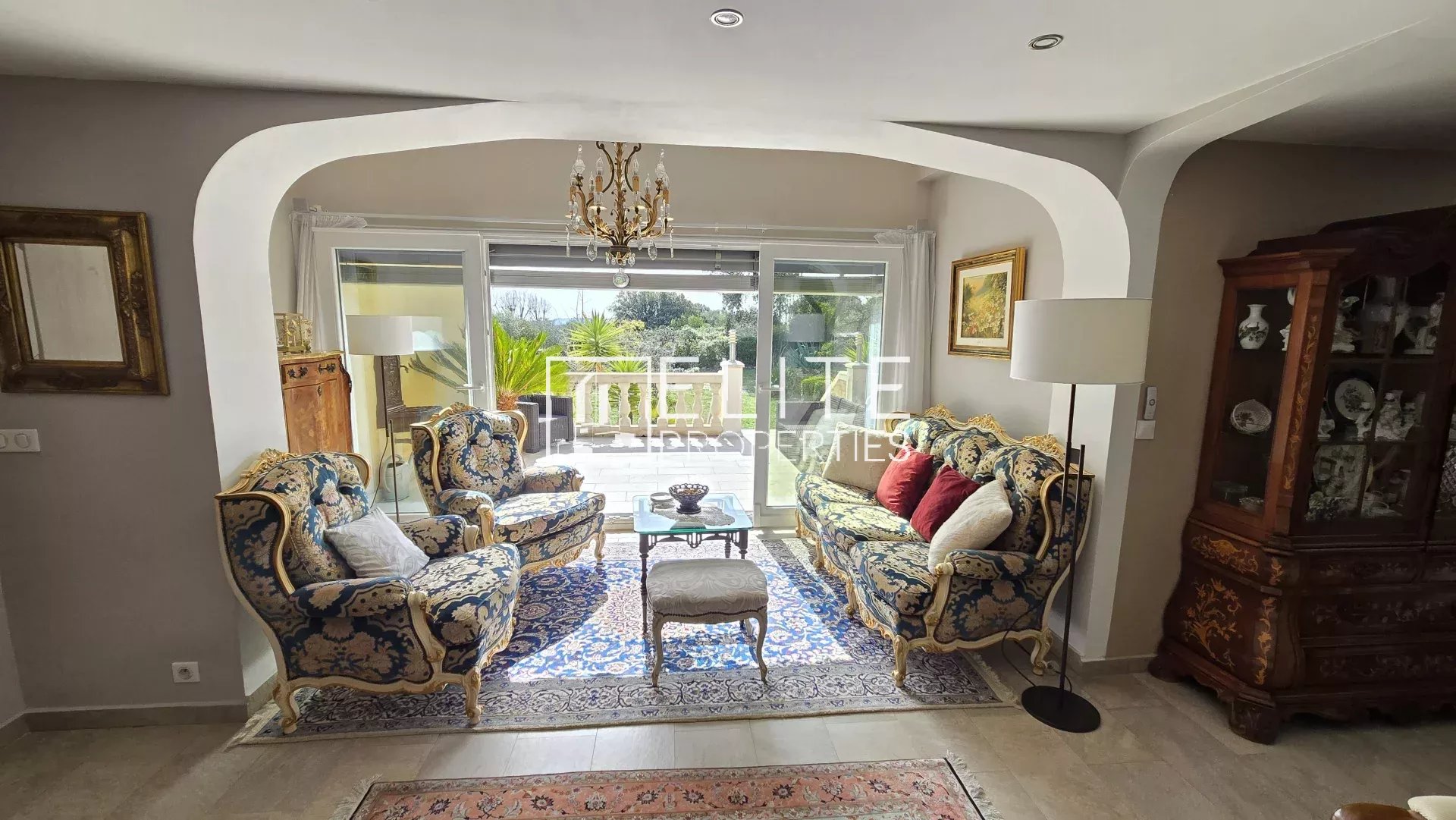 Elegant living room with blue and gold upholstered sofas facing a glass coffee table and chandelier, opening to a sunny outdoor patio through sliding doors.