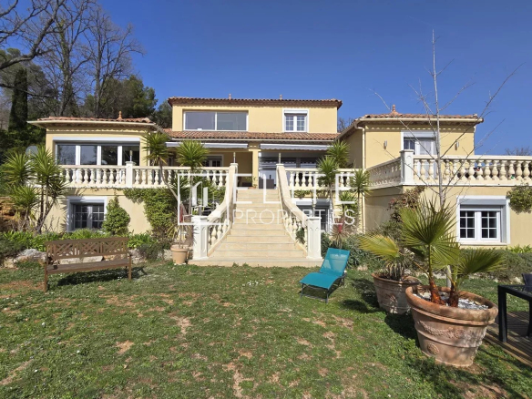 Front view of a two-story yellow villa with central staircase, white balustrades, and lush tropical landscaping on a sunny day.