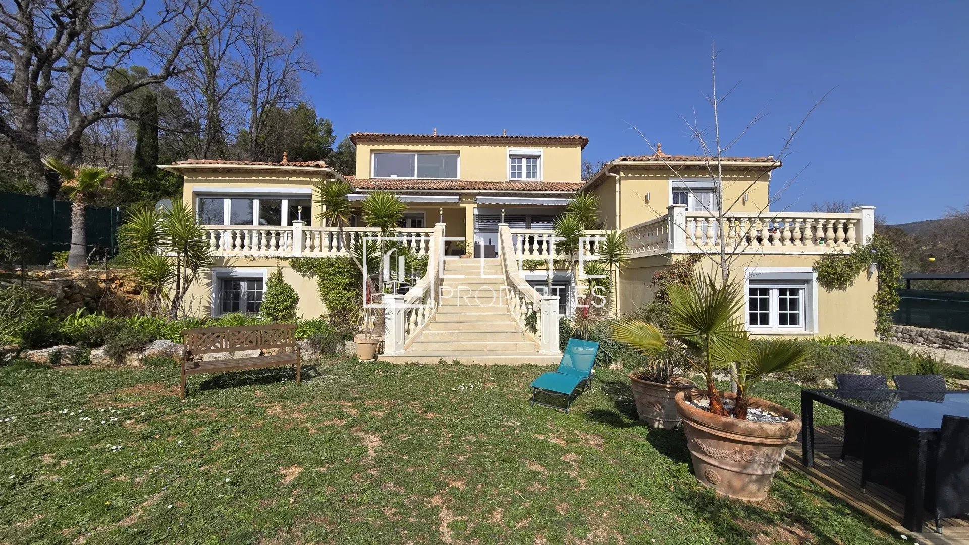 Front view of a two-story yellow villa with central staircase, white balustrades, and lush tropical landscaping on a sunny day.