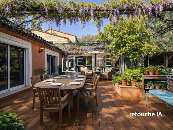 Outdoor wooden deck with a long dining table set for a meal under hanging purple wisteria, surrounded by potted plants and garden seating on a sunny day.