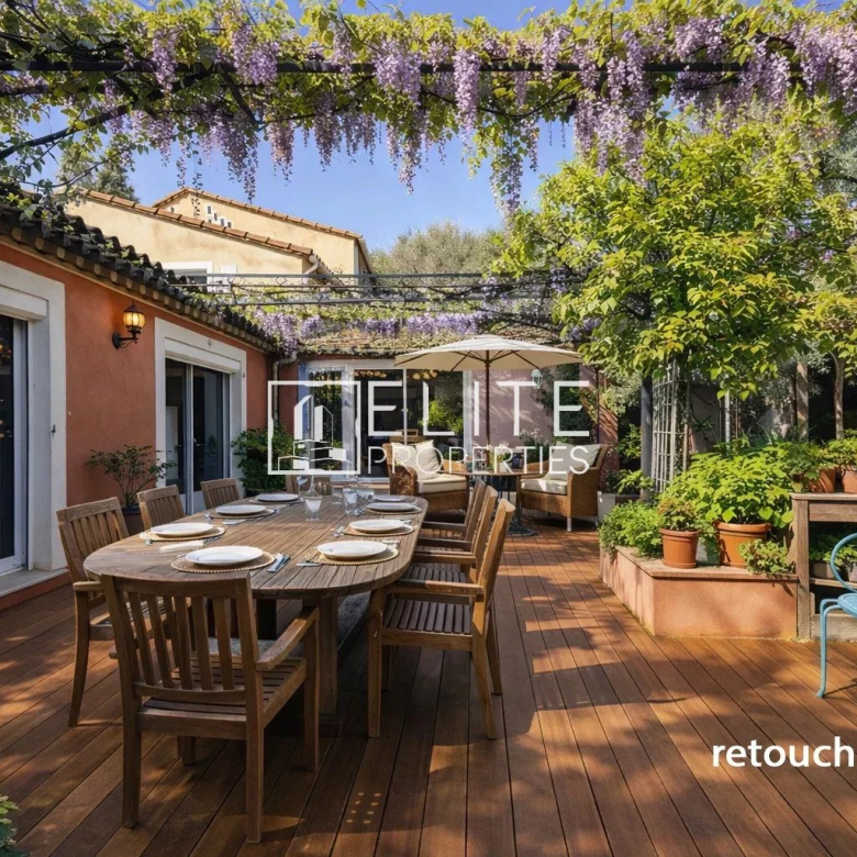 Outdoor wooden deck with a long dining table set for a meal under hanging purple wisteria, surrounded by potted plants and garden seating on a sunny day.