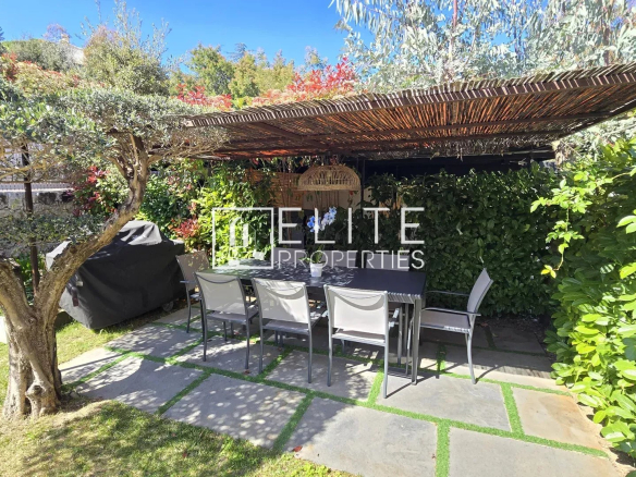 Outdoor dining area under a wooden, thatch-covered pergola with a long table and white chairs, surrounded by green hedges and trees; sunny blue sky.