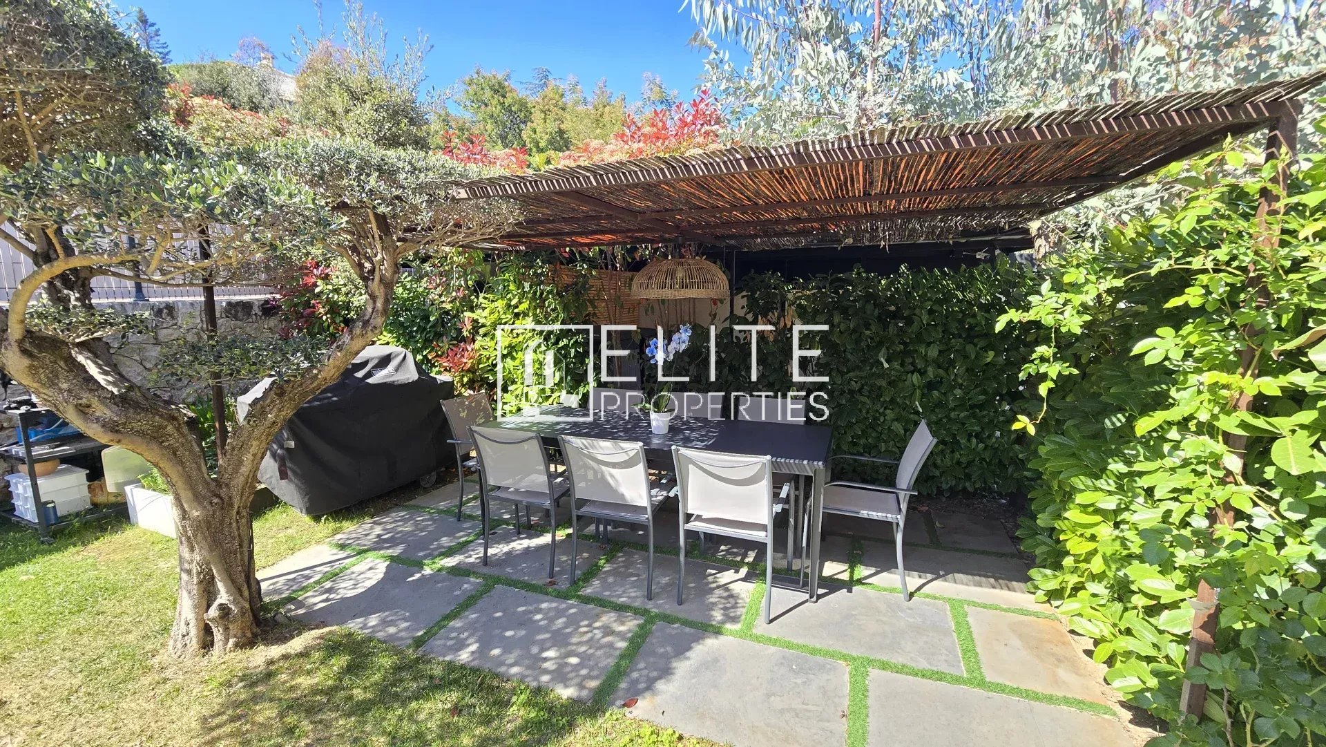 Outdoor dining area under a wooden, thatch-covered pergola with a long table and white chairs, surrounded by green hedges and trees; sunny blue sky.