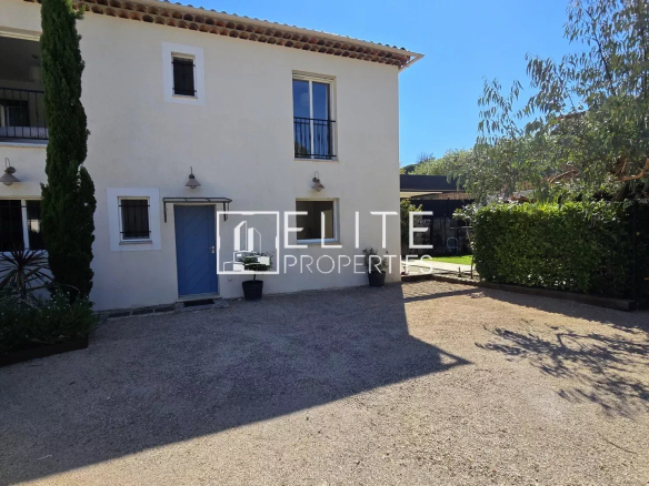 Two-story cream house with blue front door, small upper windows, and a gravel driveway amid palm-like plants and trees on a sunny day.