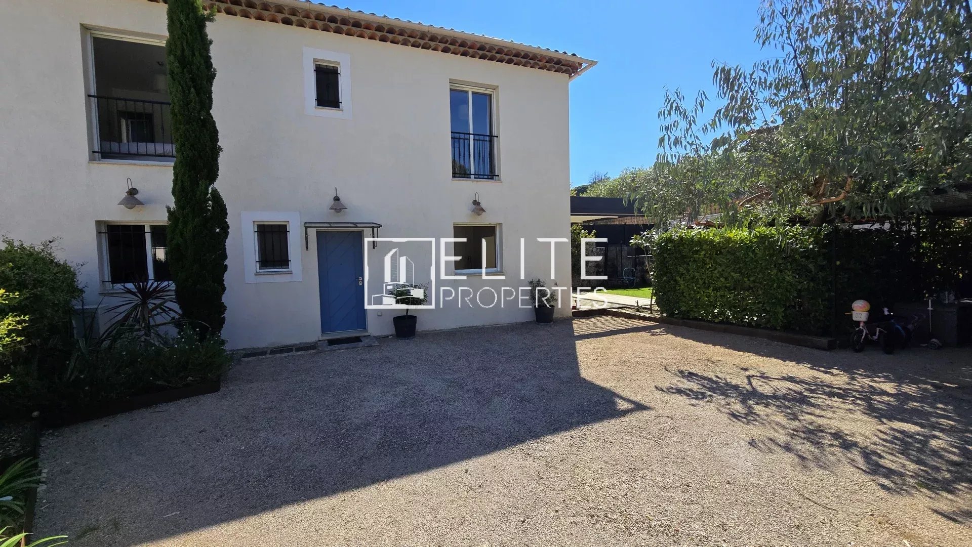 Two-story cream house with blue front door, small upper windows, and a gravel driveway amid palm-like plants and trees on a sunny day.