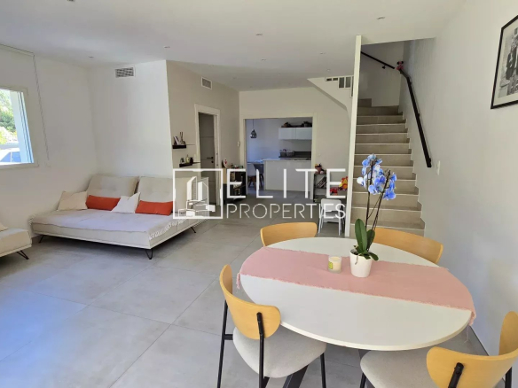 Open-plan living room with two white sofas, a round dining table with a pink table runner, and a staircase along the right wall.