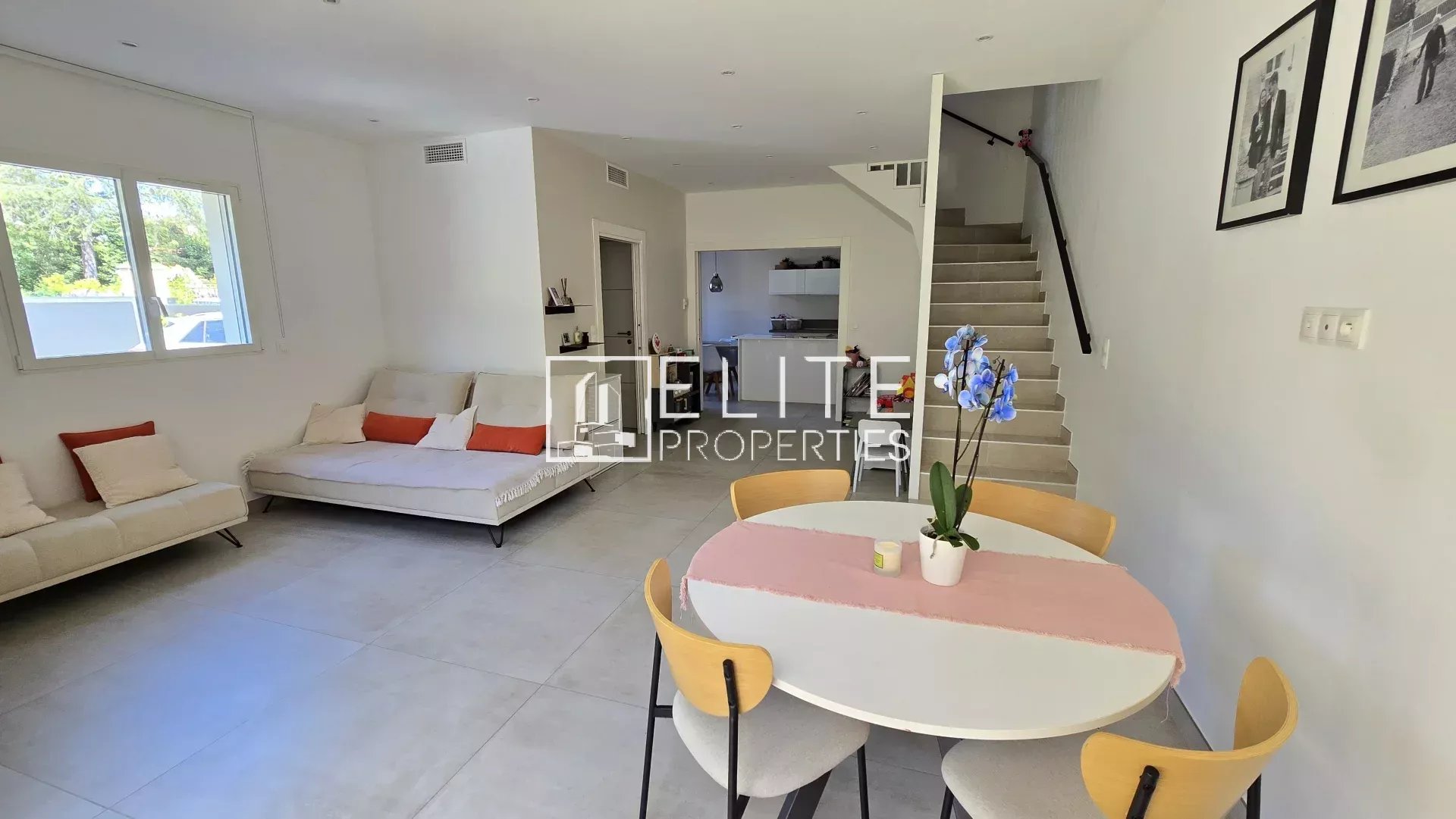 Open-plan living room with two white sofas, a round dining table with a pink table runner, and a staircase along the right wall.