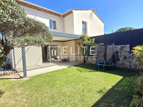 Sunlit backyard of a two-story beige house with a tiled roof, a wooden covered patio, olive tree in the foreground, and a trampoline beside a stone wall on the lawn.