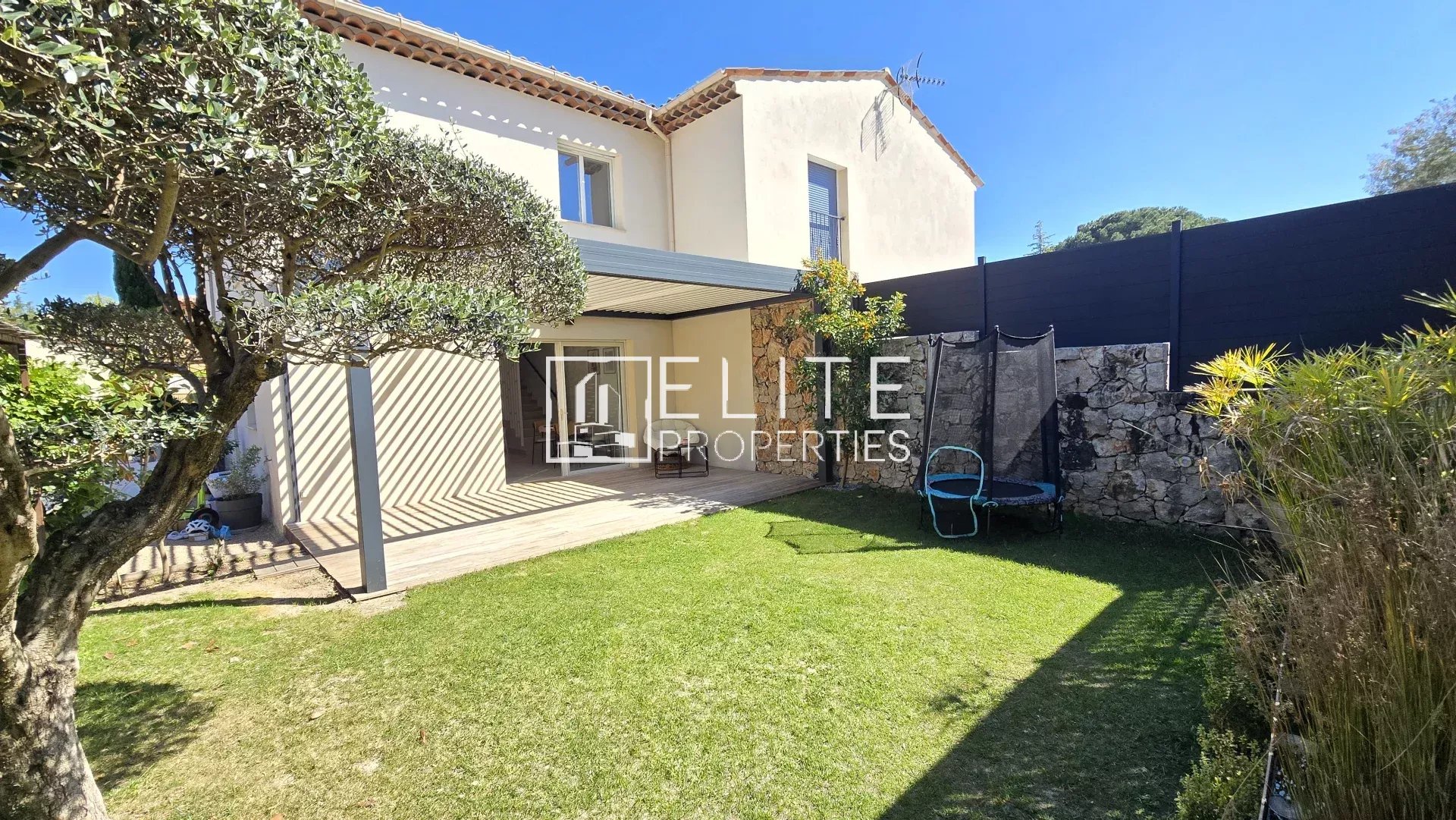 Sunlit backyard of a two-story beige house with a tiled roof, a wooden covered patio, olive tree in the foreground, and a trampoline beside a stone wall on the lawn.