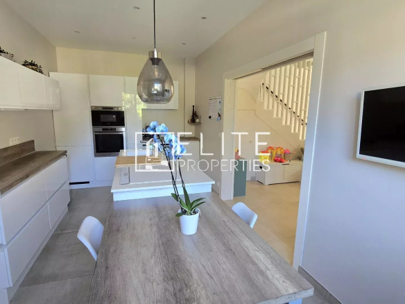 Bright modern kitchen with white cabinets and a wooden island, a glass pendant light, and a potted orchid on the table; doorway leads to a living area with stairs in the background.