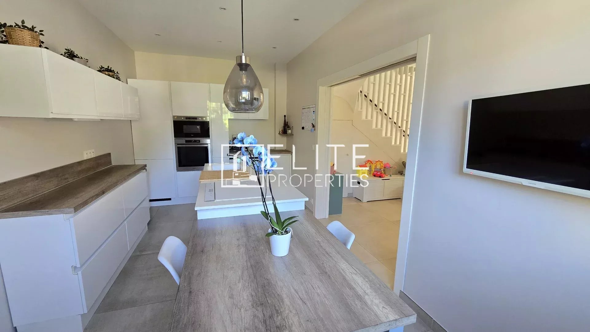 Bright modern kitchen with white cabinets and a wooden island, a glass pendant light, and a potted orchid on the table; doorway leads to a living area with stairs in the background.