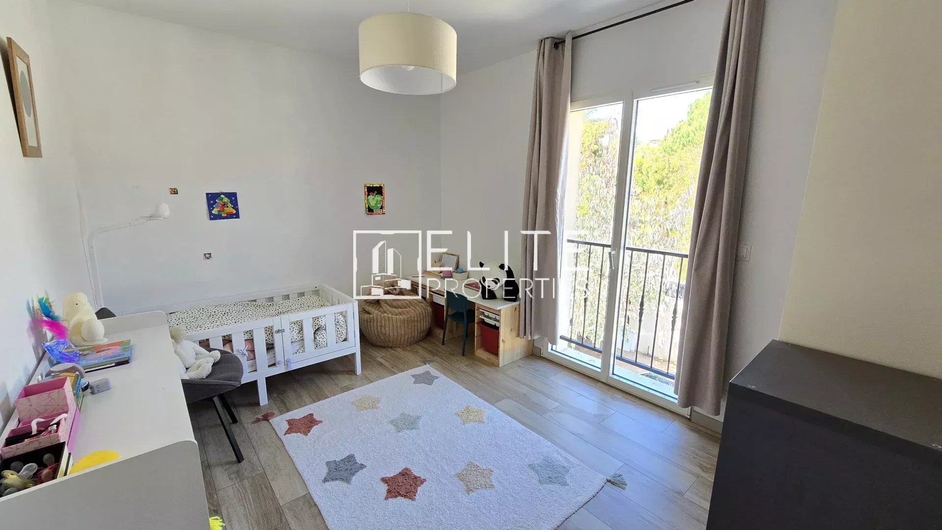 Bright child’s bedroom with a white bed, desk, and star-pattern rug beside a large window with beige curtains.
