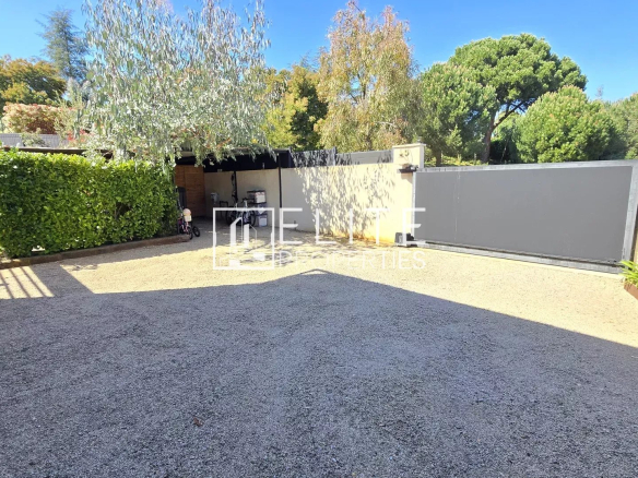 Gravel driveway with a tall hedge on the left, a covered storage area in the middle, and a large gray sliding gate on the right under a blue sky. The scene includes a bicycle and garden tools near the storage shed and greenery along the wall.