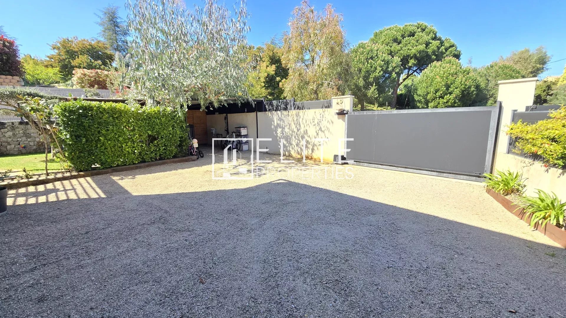 Gravel driveway with a tall hedge on the left, a covered storage area in the middle, and a large gray sliding gate on the right under a blue sky. The scene includes a bicycle and garden tools near the storage shed and greenery along the wall.