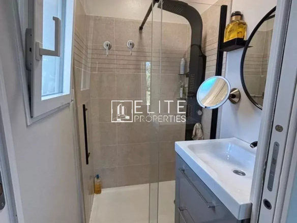 Small modern bathroom with glass‑panel shower, white sink vanity, and blue rug; window on left and round mirror on the wall.