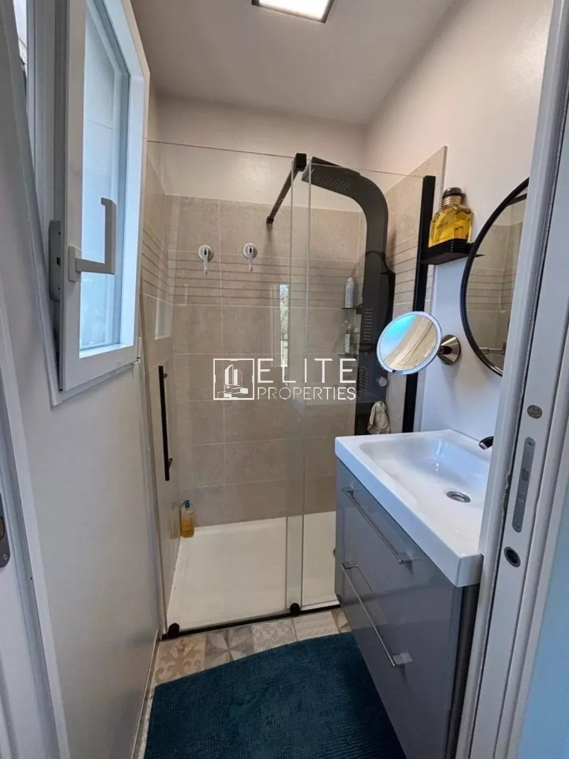 Small modern bathroom with glass‑panel shower, white sink vanity, and blue rug; window on left and round mirror on the wall.
