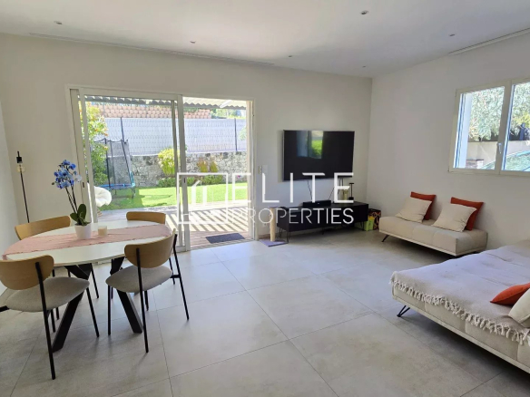 Open-plan living area with a round dining table, four wooden chairs, and a beige sofa bed facing a wall-mounted TV; sliding glass doors to a garden outside.