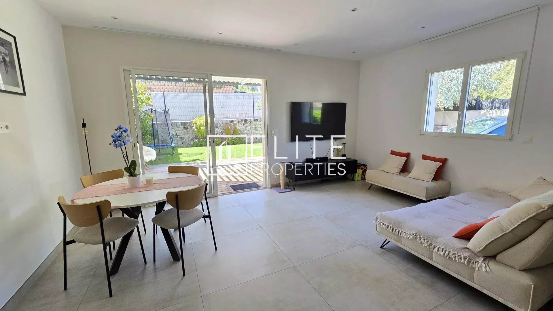 Open-plan living area with a round dining table, four wooden chairs, and a beige sofa bed facing a wall-mounted TV; sliding glass doors to a garden outside.