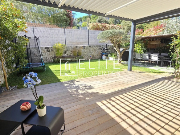 Patio area with a wooden deck under a white pergola, extending to a green lawn and stone wall, with a trampoline and outdoor dining zone beyond.