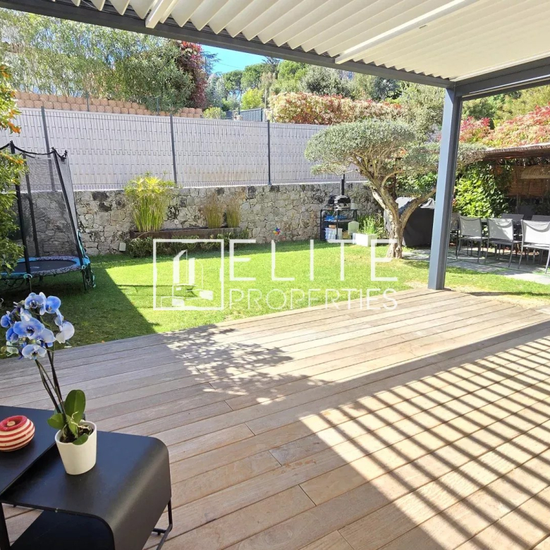 Patio area with a wooden deck under a white pergola, extending to a green lawn and stone wall, with a trampoline and outdoor dining zone beyond.