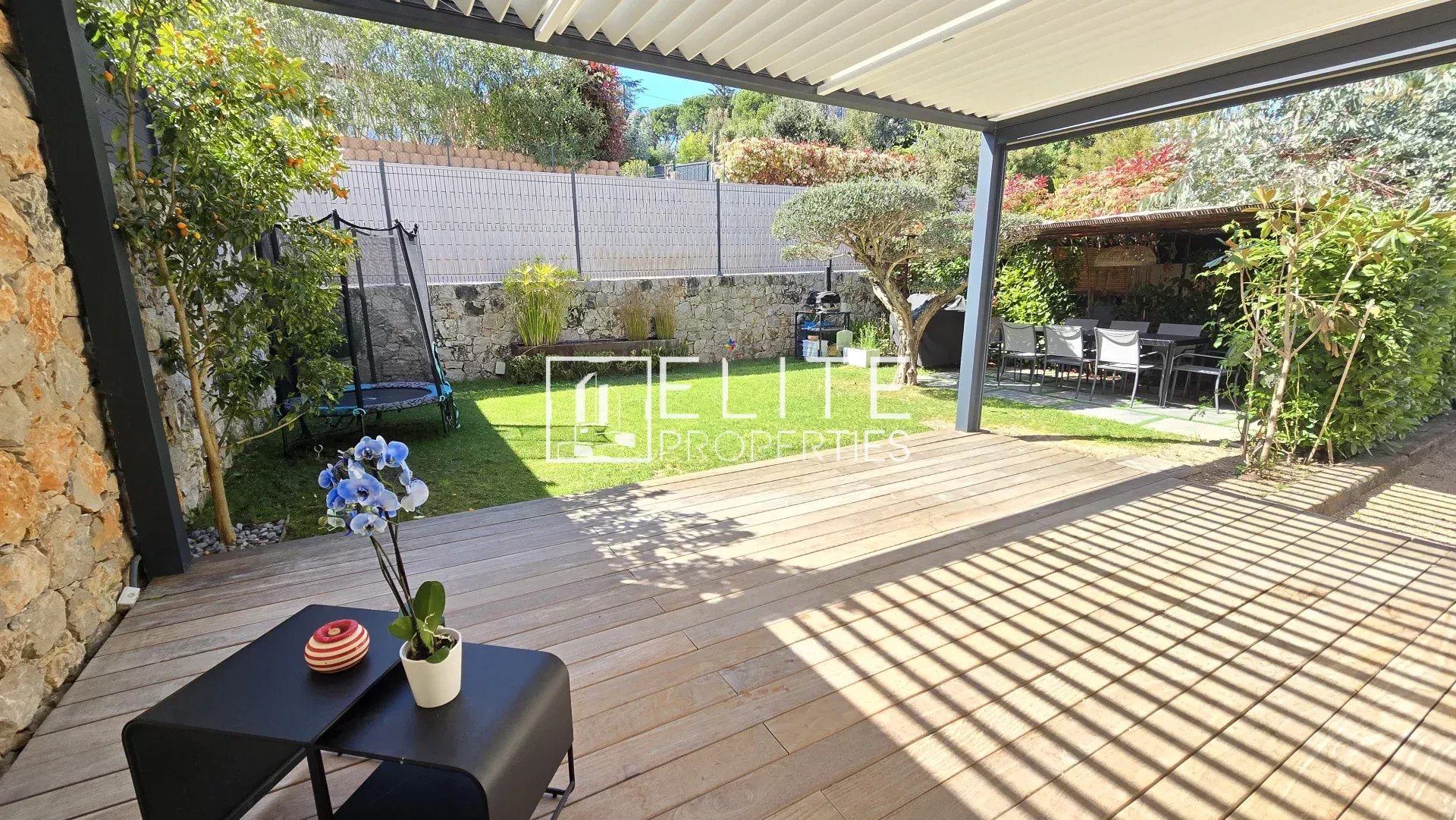 Patio area with a wooden deck under a white pergola, extending to a green lawn and stone wall, with a trampoline and outdoor dining zone beyond.