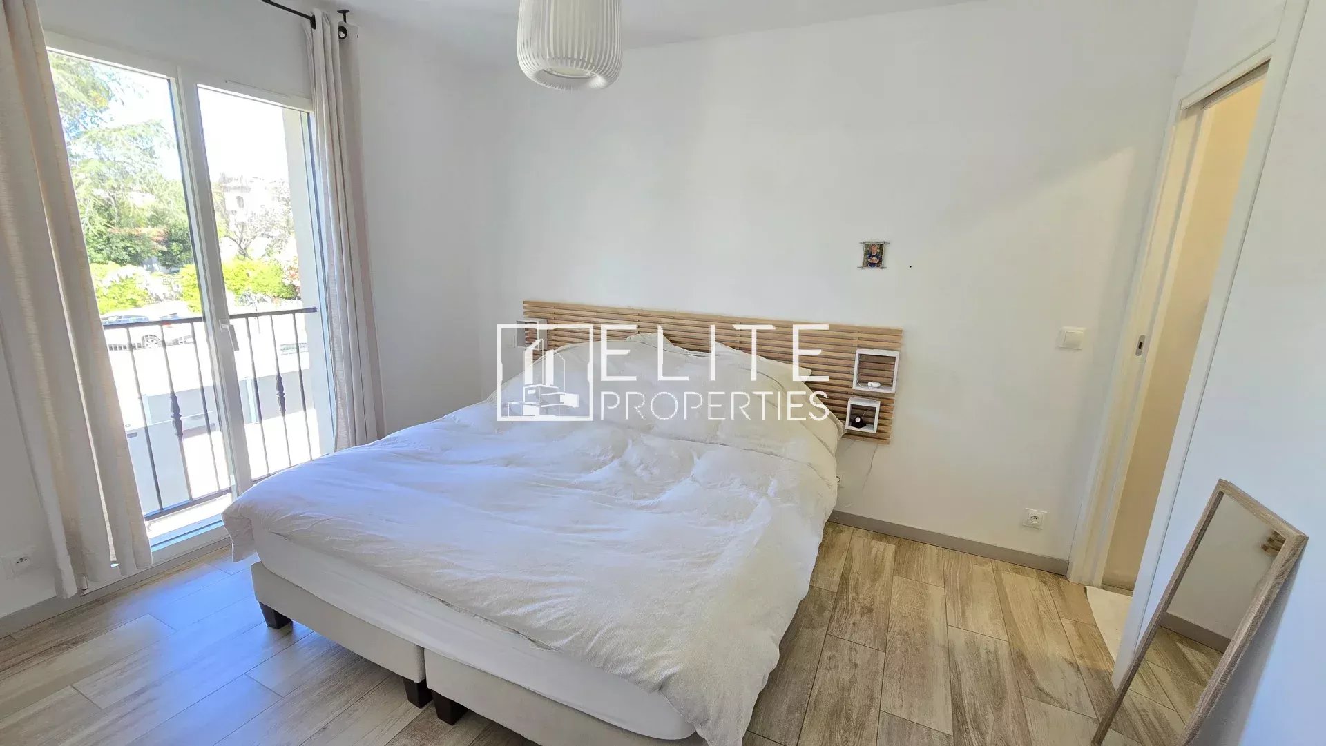 Bright bedroom with a double bed, white bedding, and a wooden slatted headboard near a balcony door.