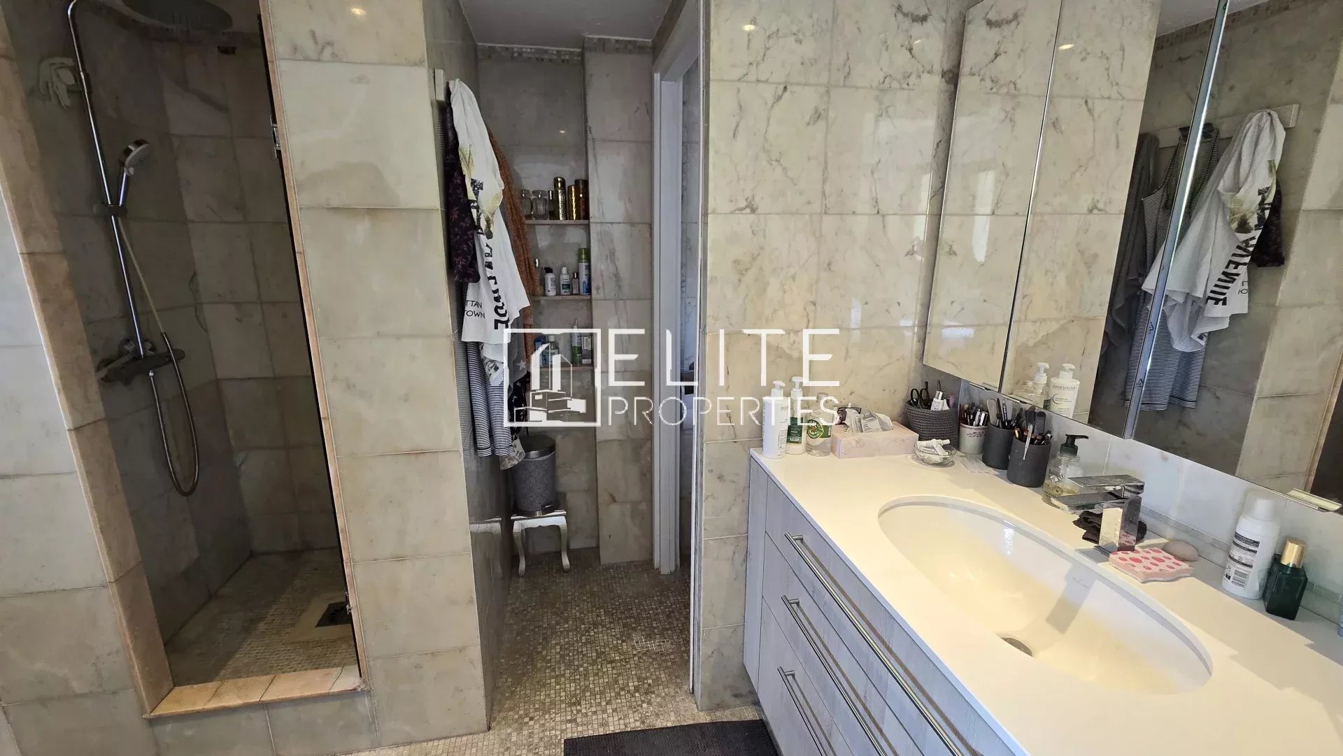 Salle de bain moderne avec douche et lavabo, murs en marbre, produits de beauté sur le plan de travail.
