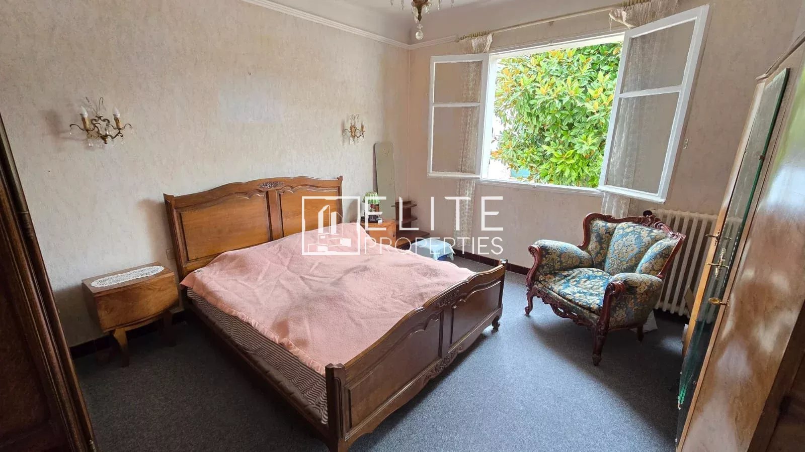 Chambre d’appartement à Juan-les-Pins Chambre d'un appartement avec un lit en bois et un fauteuil, fenêtres ouvertes sur l'extérieur.