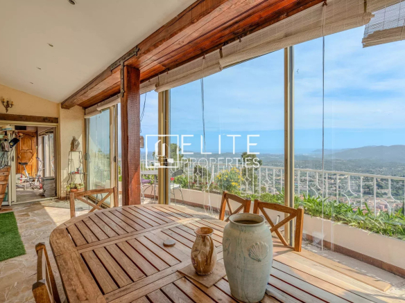 Vue panoramique depuis une véranda avec table en bois et décoration, surplombant la mer et la ville de Grasse.