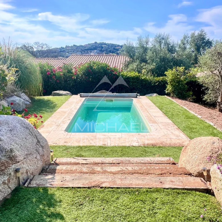 Residential backyard with a rectangular turquoise pool, stone surround, green lawn, and shrubs along a hedge under a blue sky.