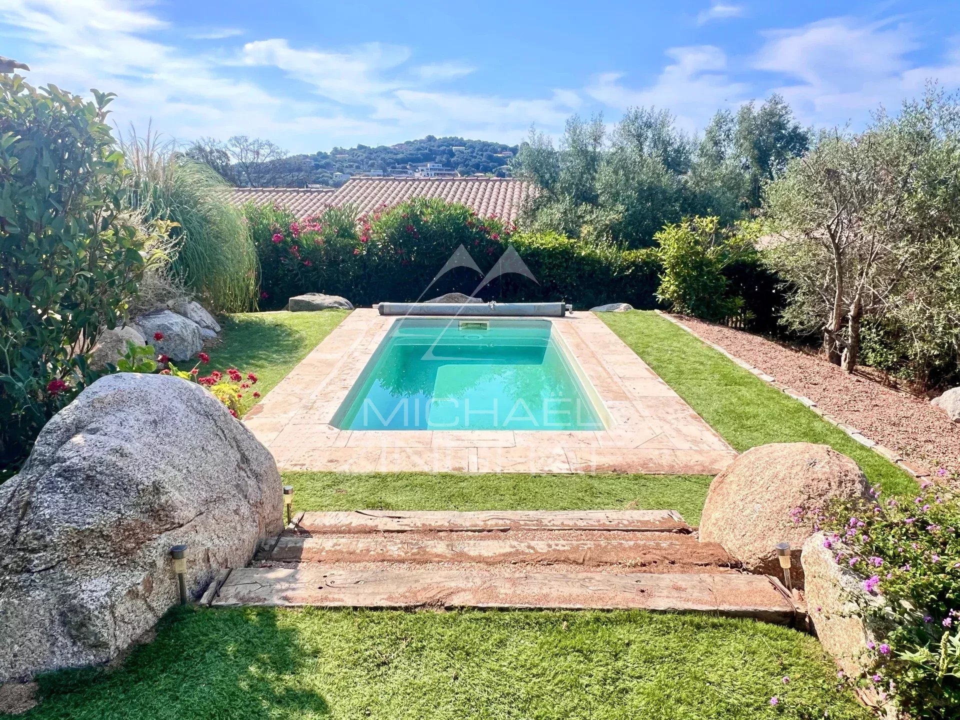 Residential backyard with a rectangular turquoise pool, stone surround, green lawn, and shrubs along a hedge under a blue sky.