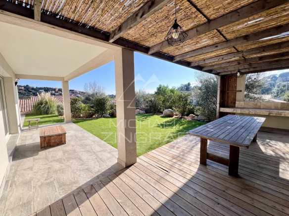 Covered patio with a wooden deck and table overlooking a green backyard and hillside village on a sunny day