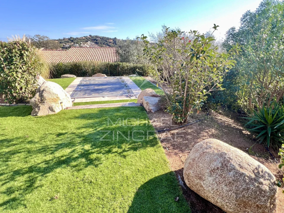 Sunlit backyard garden with a rectangular pool covered by a safety cover, surrounded by rocks, green lawn, and shrubs.