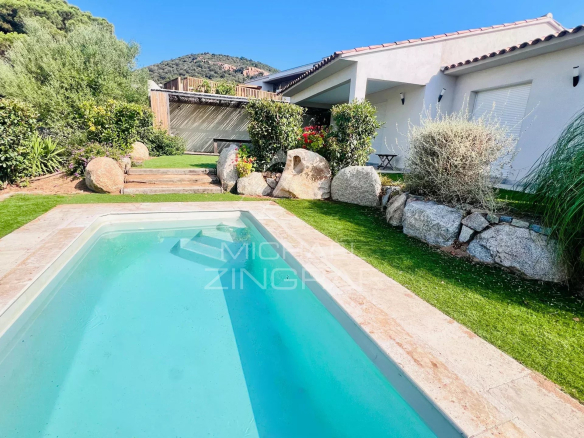 Backyard with a rectangular in-ground pool, white house, stone border, and lush green lawn under a clear blue sky.