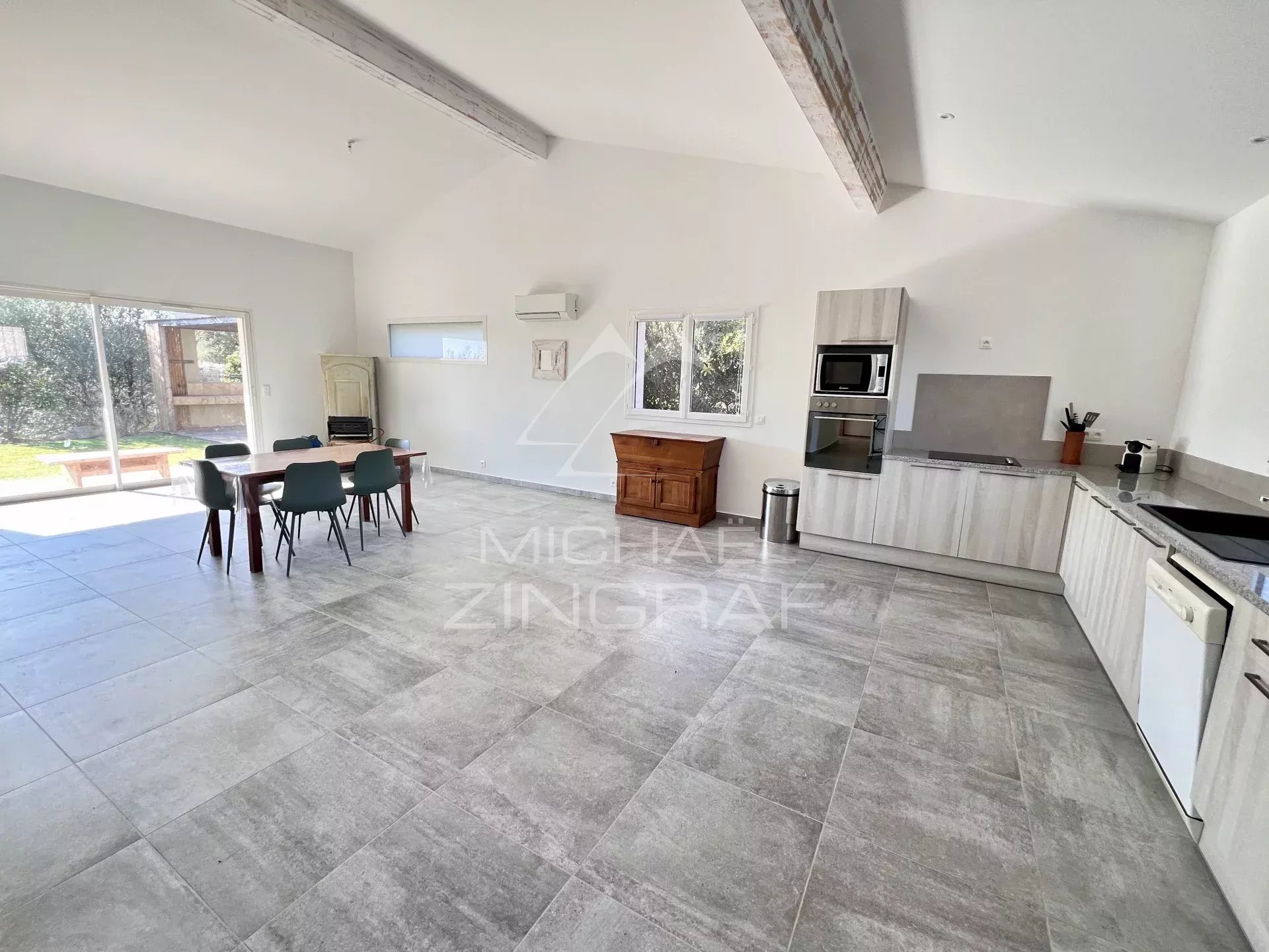 Open-plan kitchen and dining area with a wooden table and green chairs, sliding glass doors to a backyard, and light gray cabinetry with built-in appliances.