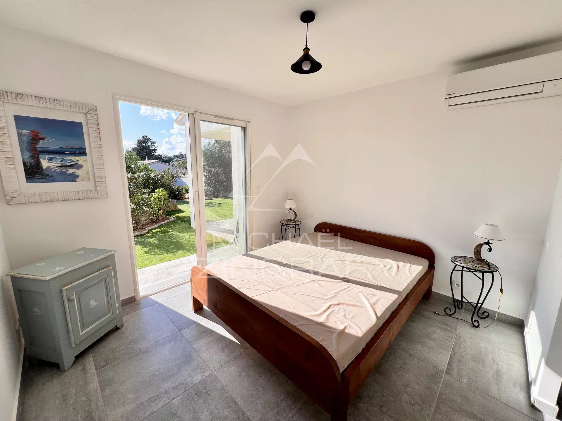 Bright bedroom with a wooden bed frame, two small metal bedside tables with lamps, and a sliding glass door opening to a green garden view.