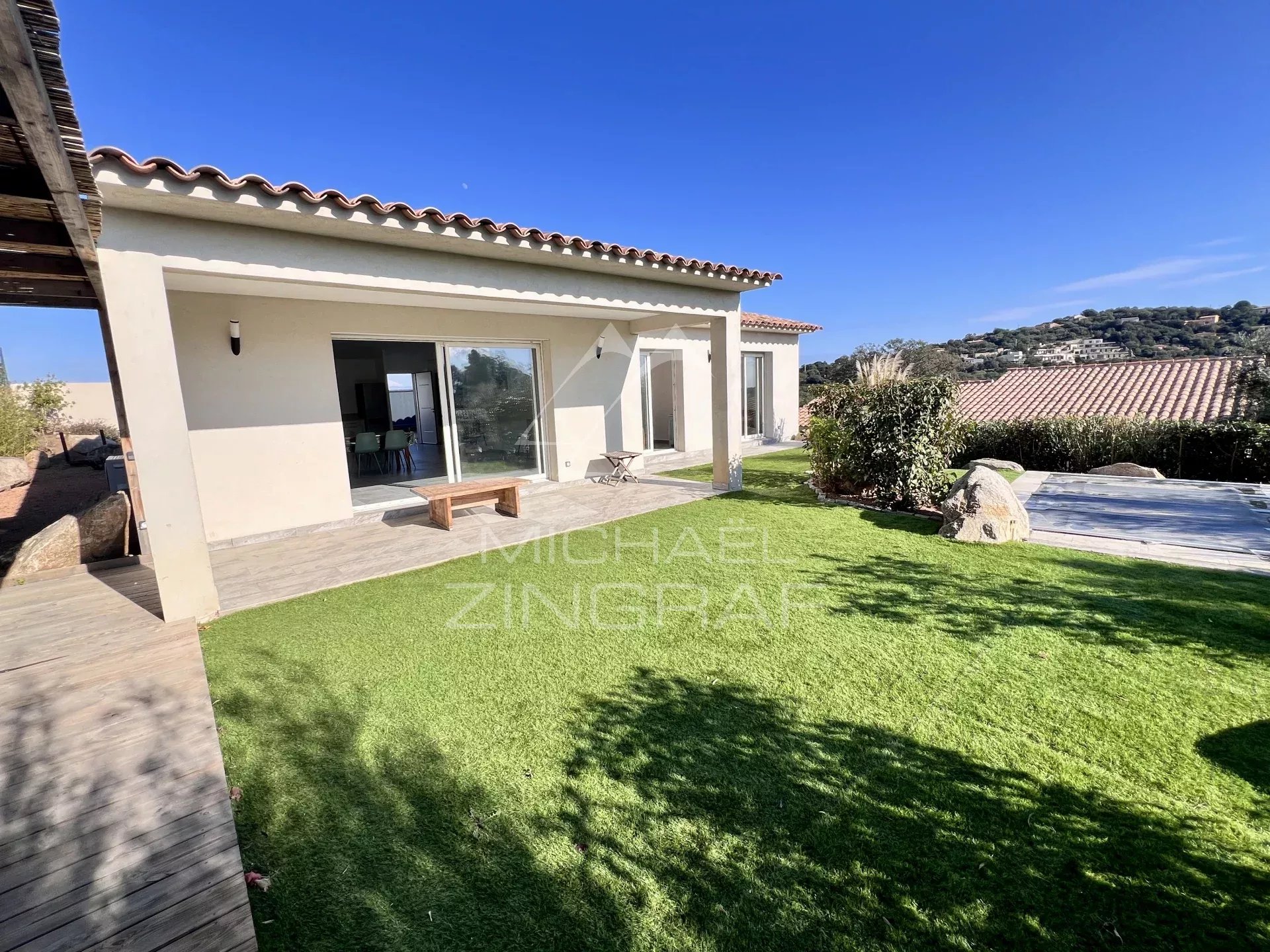 Single-story house with beige exterior and terracotta roof, sliding glass doors open to a patio overlooking a green lawn.