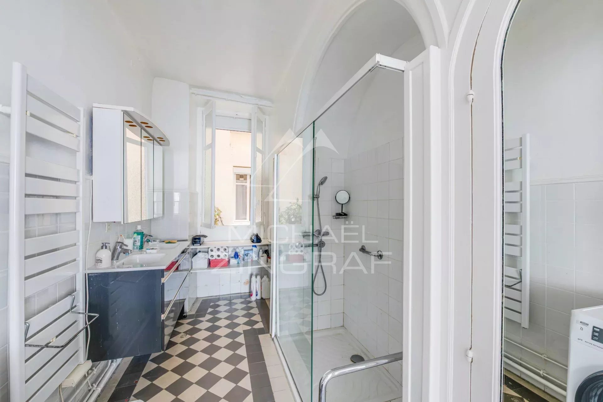 Salle de bain moderne avec douche et lavabo, carrelage noir et blanc, fenêtre laissant entrer la lumière.
