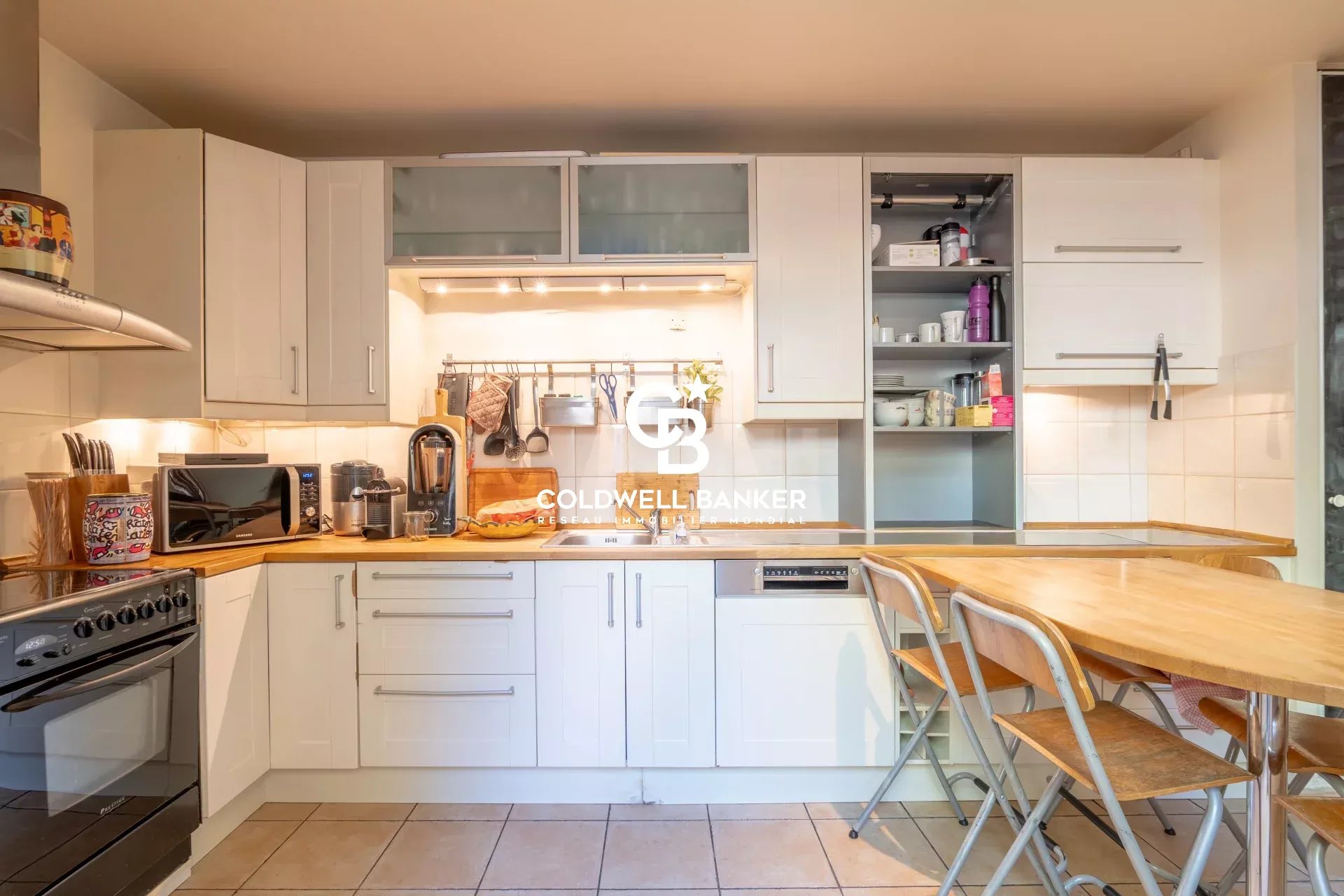 Bright white kitchen with wooden countertops, appliances, and open-shelving pantry; a wooden dining table with metal chairs on the right.