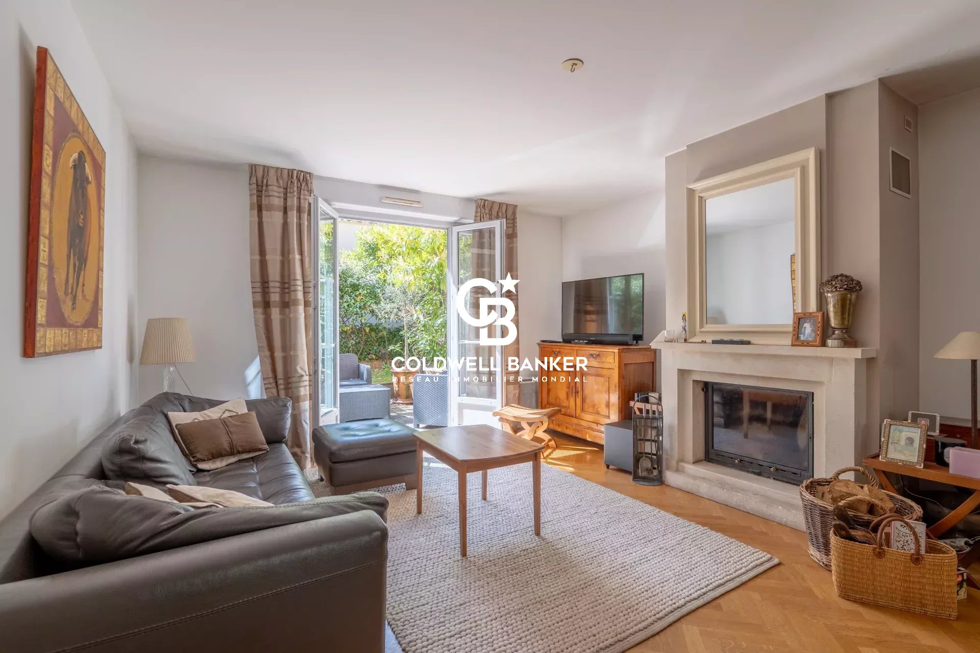 Living room with a gray leather sofa and matching ottoman facing a fireplace and TV, bright French doors open to a garden patio outside.