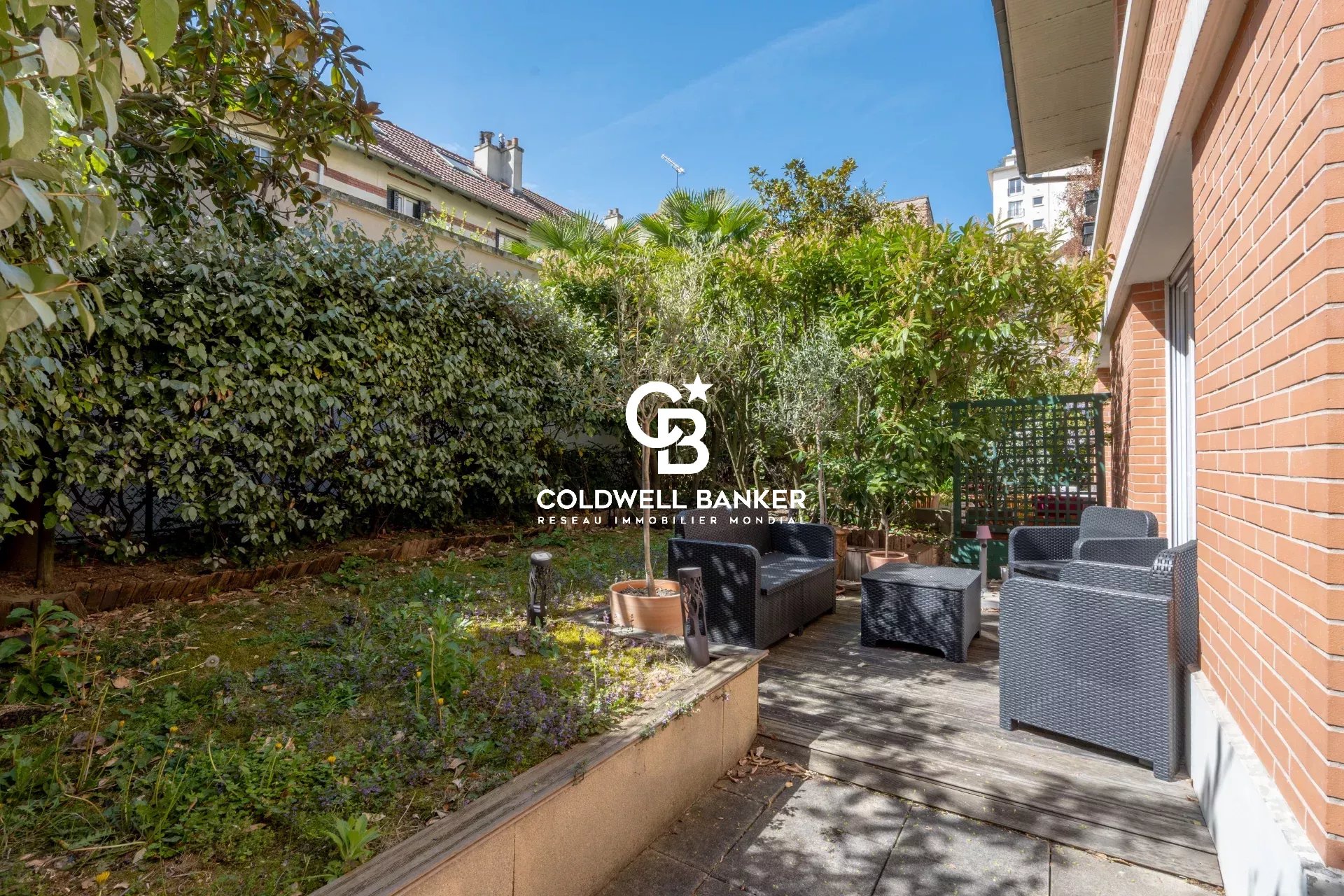 Urban garden patio with wicker seating and potted trees along a brick house, logo of Coldwell Banker centered in the scene.