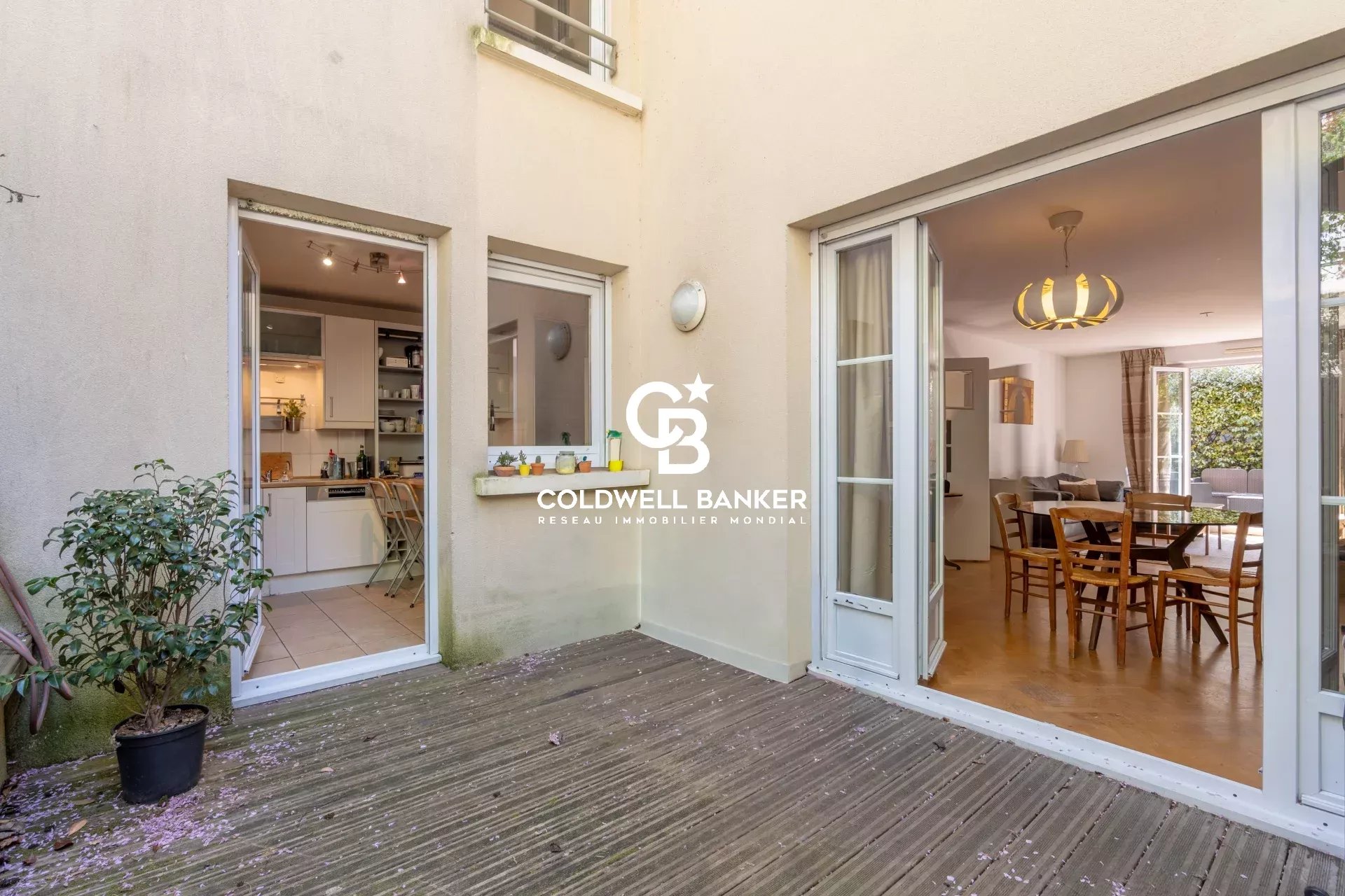 Patio with a wooden deck and two open doors: left shows a white kitchen, right opens to a dining room; Coldwell Banker branding on the wall between.