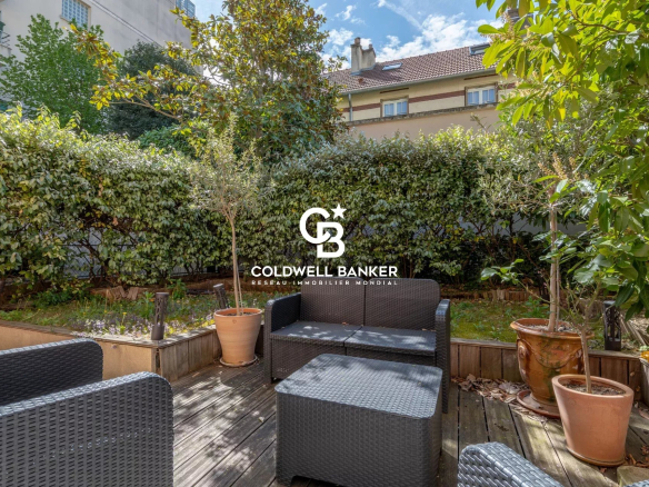 Sunny outdoor patio with black wicker furniture on a wooden deck, potted plants, and a tall hedge backdrop, under a blue sky.